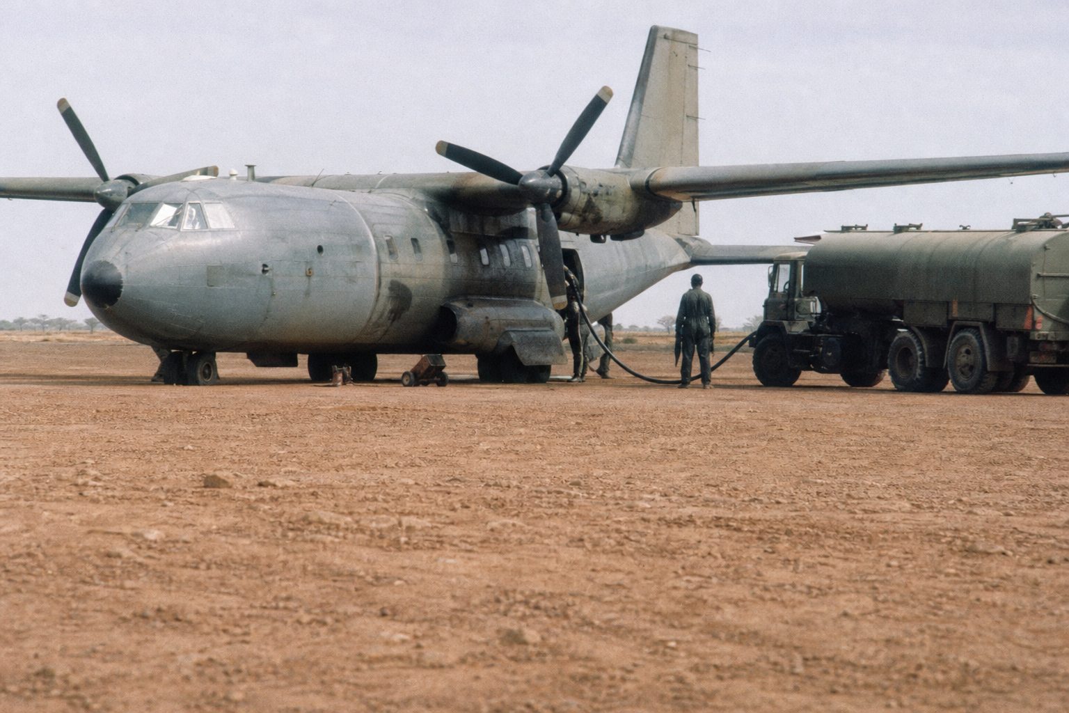 Ageing twin-propeller military transport plane on a hot runway with a fuel truck alongside (picture made with AI)