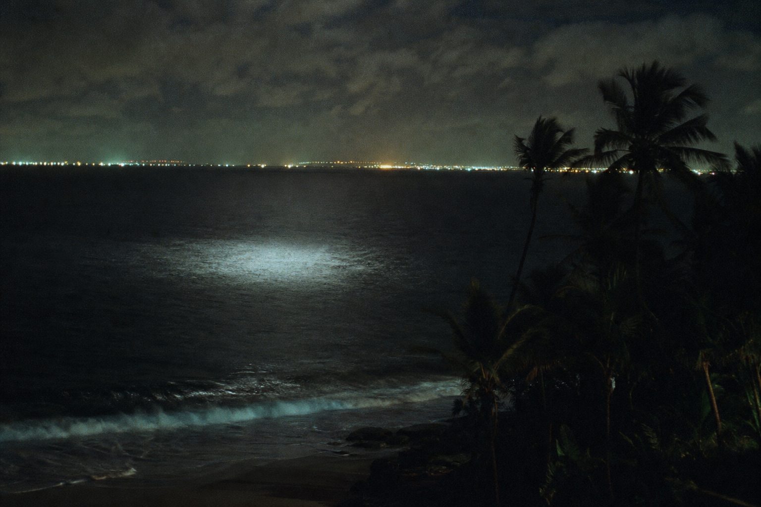 Dark sea at night off a tropical coast with distant city lights on the horizon (picture made with AI)