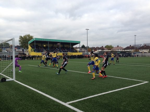 Players in yellow and blue and in black challenge for a corner on an artificial pitch