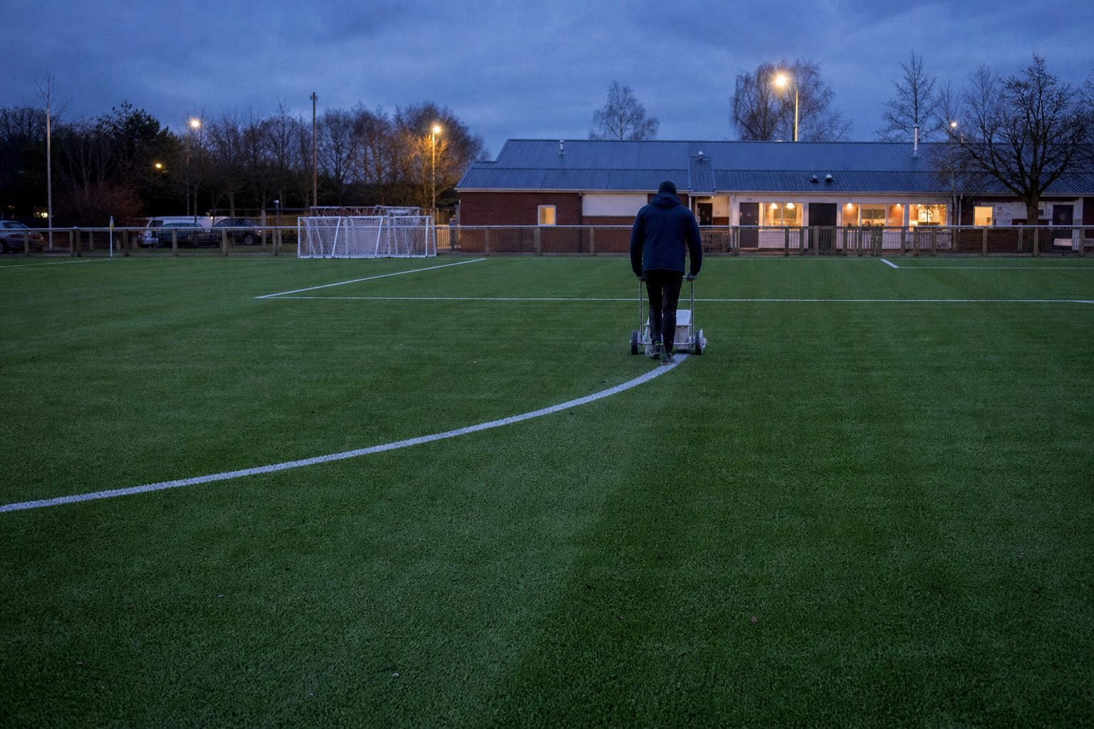 A groundsman marking out a small artificial pitch at a community club at dusk (picture made with AI)