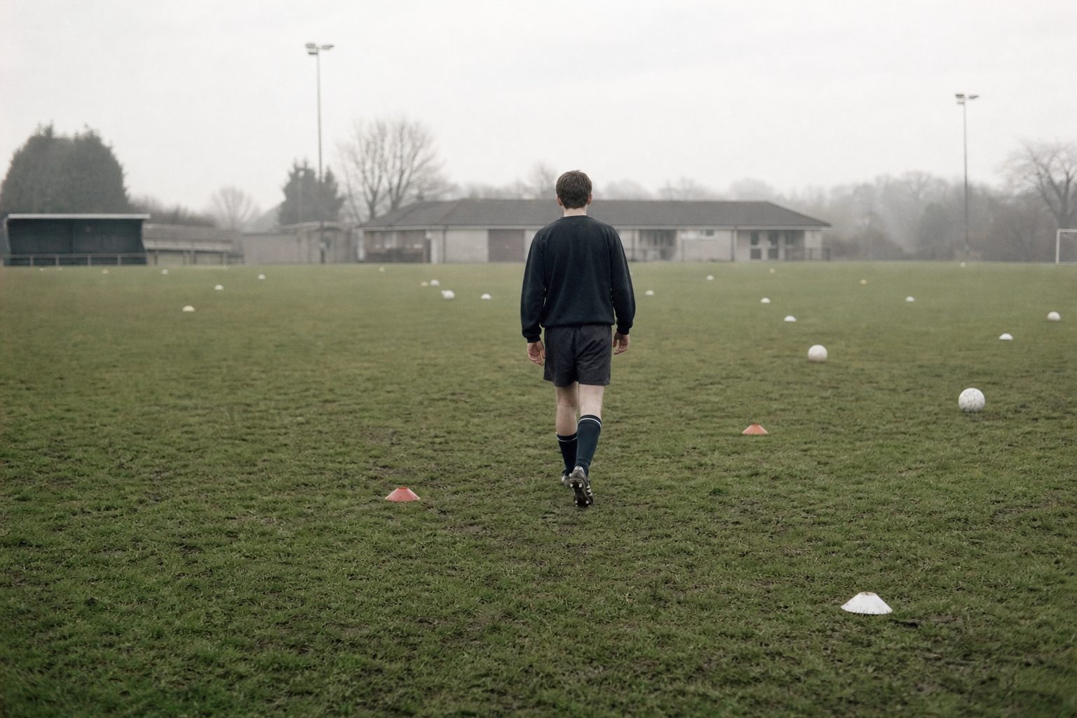 Young player seen from behind walking across an empty misty training pitch (picture made with AI)