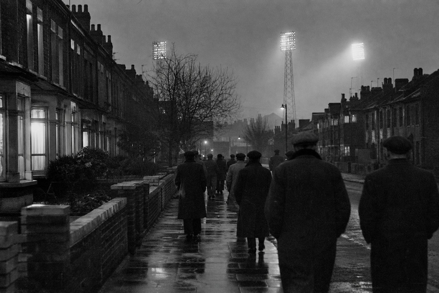 Men in overcoats walking down a wet terraced street towards distant floodlight pylons at dusk (picture made with AI)