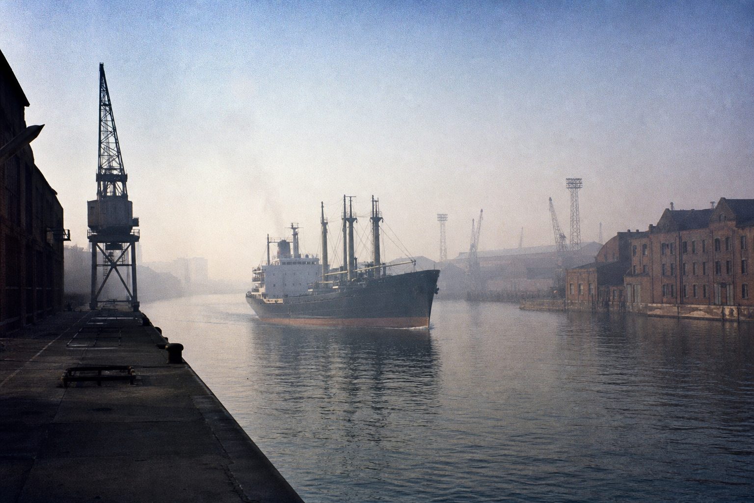 Cargo ship moving along a misty industrial canal between brick warehouses and cranes (picture made with AI)