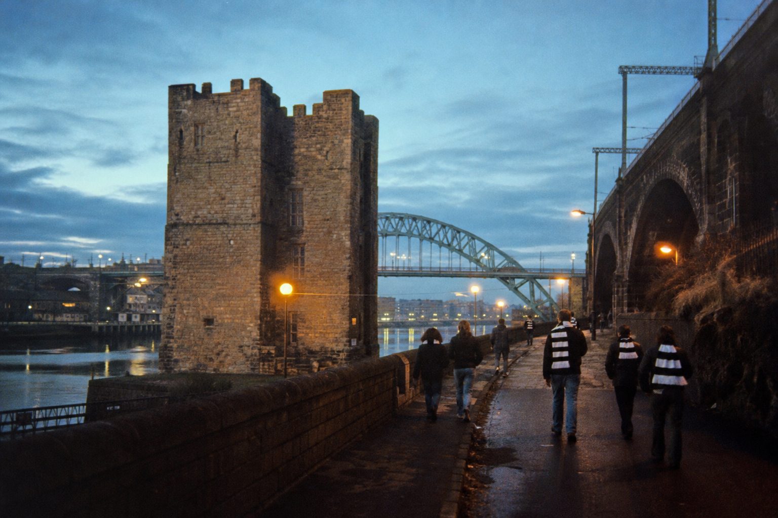 Stone castle keep beside railway arches over a river at dusk, fans walking uphill (picture made with AI)