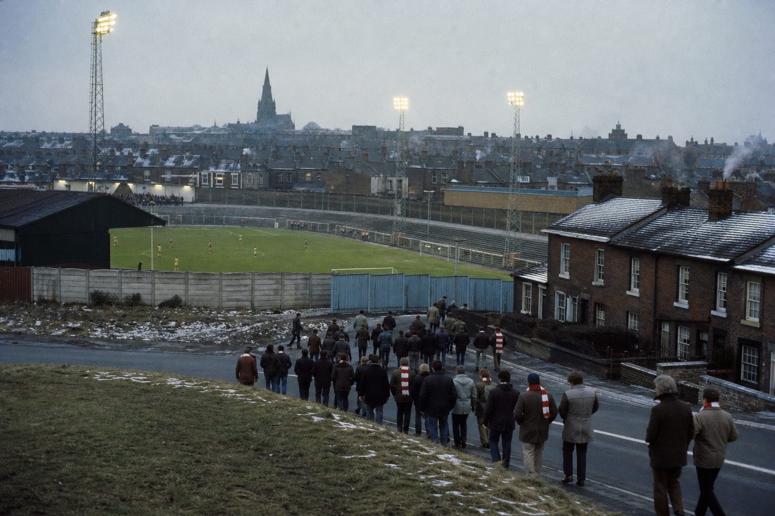 Old football ground among terraced houses at dusk with floodlights just switched on (picture made with AI)
