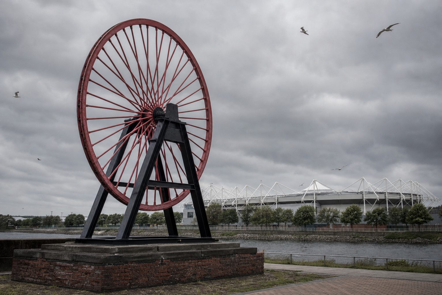 Preserved colliery winding wheel on a plinth beside a riverside stadium under grey skies (picture made with AI)