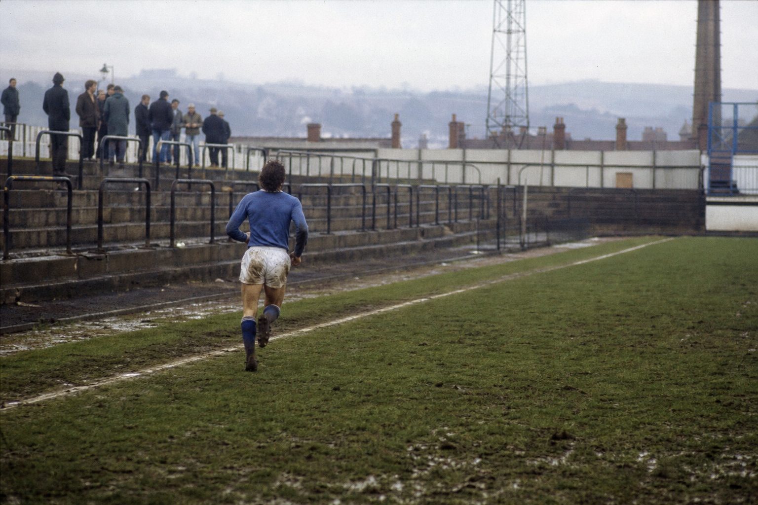 Lone full-back sprinting down the touchline of a muddy pitch in front of an old terrace (picture made with AI)