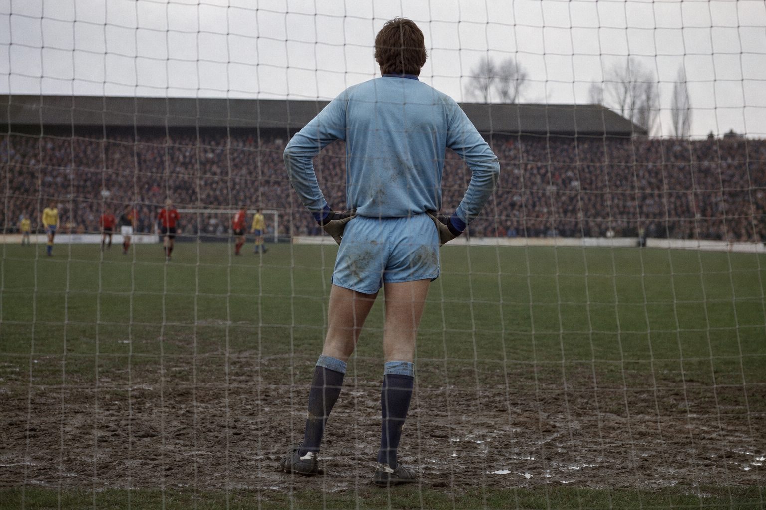 Tall goalkeeper seen from behind on his goal line in front of a packed terrace (picture made with AI)
