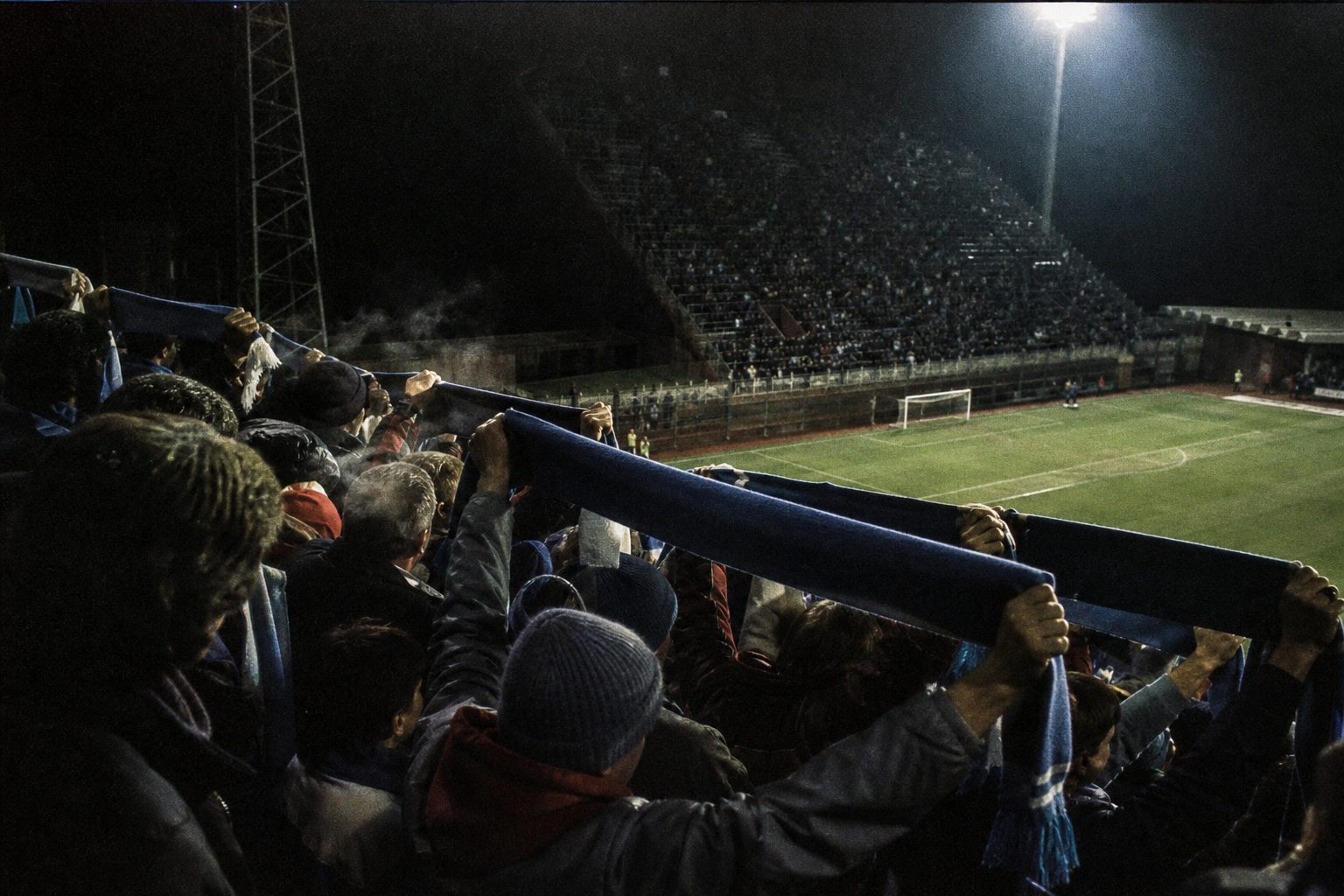 Travelling supporters with raised scarves in a packed away end on a cold floodlit night (picture made with AI)